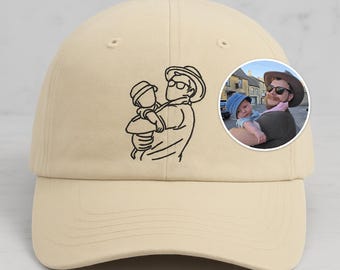Könnte beinhalten: Beige Baseball Cap mit einer schwarzen Strichzeichnung einer Person, die ein Kind hält. Auf der Seite befindet sich ein rundes Foto einer Person, die ein Baby hält. Die Kappe hat einen gebogenen Schirm und einen verstellbaren Riemen.