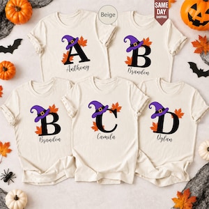 Könnte beinhalten: Fünf beige T-Shirts mit kurzen Ärmeln, verziert mit einem großen schwarzen Buchstaben, einem violetten Hexenhut und orangefarbenen Herbstblättern. Jedes T-Shirt zeigt einen Namen in schwarzer Schreibschrift: Anthony, Brandon, Camila oder Dylan.