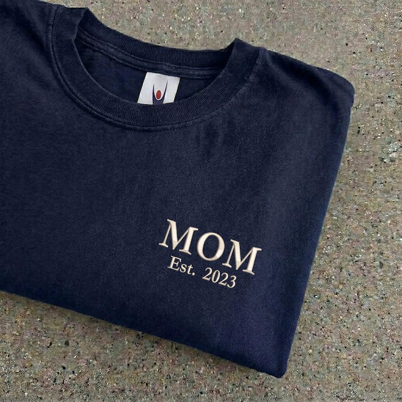 May include: Navy blue crew neck t-shirt with the word "MOM" embroidered in a cream color, along with "Est. 2023". The shirt has a classic, casual style.