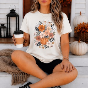 以下が含まれることがあります： クリーム色の半袖Tシャツで、中央にカボチャと、オレンジ、茶色、落ち着いた緑色の秋らしい花や葉が描かれたグラフィックプリントが施されており、黒いショートパンツと組み合わされています。