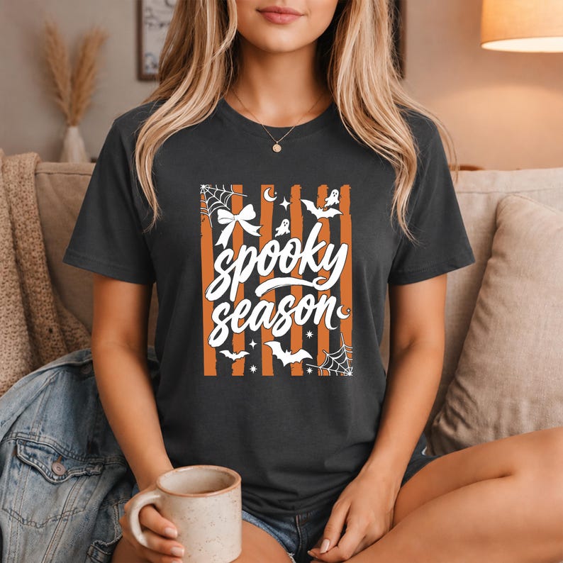 May include: A dark grey short-sleeve t-shirt featuring a graphic print with vertical orange and black stripes. The design includes the white text "spooky season" surrounded by white illustrations of a bow, spiderwebs, bats, ghosts, and stars.