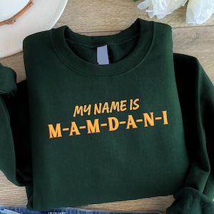 May include: Dark green sweatshirt with the text "MY NAME IS M-A-M-D-A-N-I" in orange. The sweatshirt is folded, with the sleeves visible. A white hat and a flower are in the background.