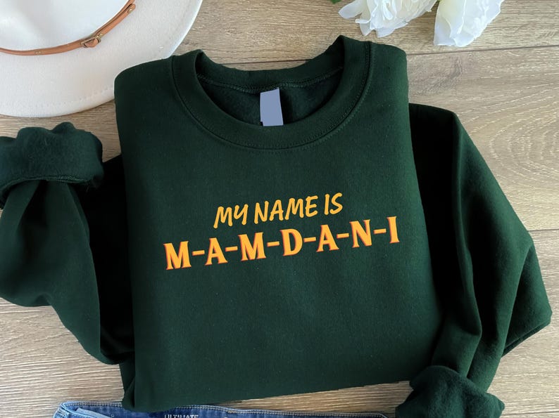 May include: Dark green sweatshirt with the text "MY NAME IS M-A-M-D-A-N-I" in orange. The sweatshirt is folded, with the sleeves visible. A white hat and a flower are in the background.
