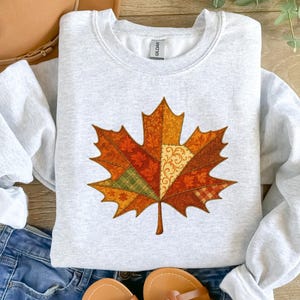Könnte beinhalten: Ein hellgraues Sweatshirt mit einem großen, farbenfrohen Ahornblatt-Design. Das Blatt besteht aus verschiedenen herbstlichen Mustern in Orange-, Rot-, Grün- und Gelbtönen. Das Sweatshirt wird mit einer blauen Jeans und Sandalen kombiniert.