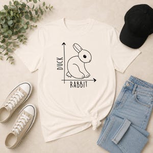 Może przedstawiać: Kremowy t-shirt z krótkim rękawem z czarnym rysunkiem liniowym królika umieszczonym na wykresie. Oś pionowa jest oznaczona jako DUCK, a oś pozioma jako RABBIT. Wzór znajduje się na środku przedniej części koszulki.