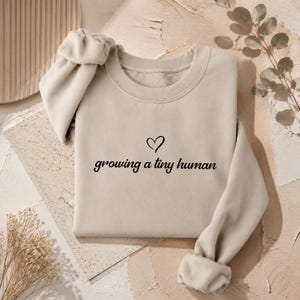 May include: A beige crewneck sweatshirt featuring the black text "growing a tiny human" printed on the centre chest, accompanied by a small black heart outline positioned directly above the text. The garment has long sleeves with ribbed cuffs.