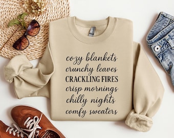 May include: A beige sweatshirt with the words "cozy blankets, crunchy leaves, crackling fires, crisp mornings, chilly nights, comfy sweaters" in black script. The sweatshirt is next to brown shoes, sunglasses, and denim jeans.
