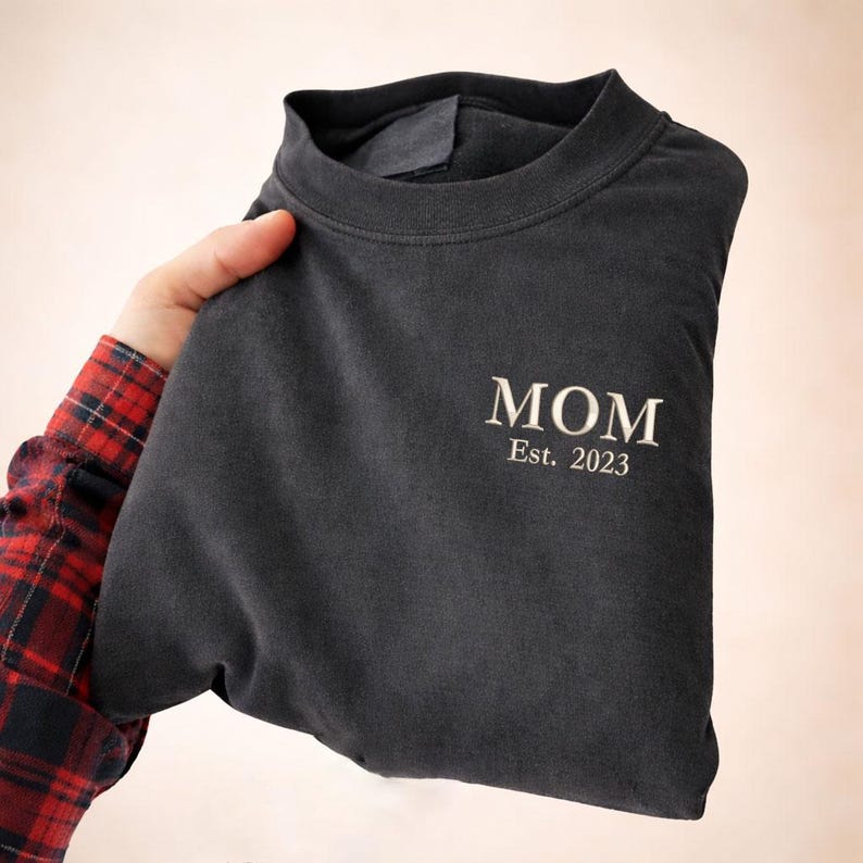 May include: A dark gray sweatshirt with the word "MOM" embroidered in white, along with "Est. 2023". The sweatshirt is folded and held by a person wearing a red and black plaid shirt.