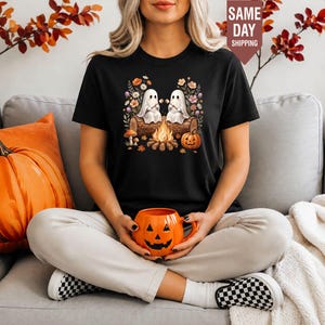May include: A black t-shirt featuring a graphic of two white ghosts sitting on a log by a campfire, surrounded by colourful flowers and a pumpkin. The ghosts are roasting marshmallows. The shirt is held by someone holding an orange pumpkin mug.