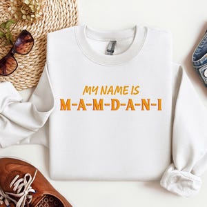 May include: A white sweatshirt with the text "MY NAME IS M-A-M-D-A-N-I" in orange and yellow lettering. The sweatshirt is displayed with brown shoes, sunglasses, and a pair of blue jeans.