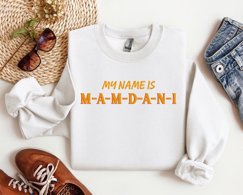 May include: A white sweatshirt with the text "MY NAME IS M-A-M-D-A-N-I" in orange and yellow lettering. The sweatshirt is displayed with brown shoes, sunglasses, and a pair of blue jeans.