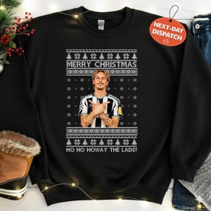 Puede incluir: Sudadera negra con un diseño navideño festivo. El diseño incluye el texto "MERRY CHRISTMAS" y "HO HO HOWAY THE LADS!" junto con patrones de copos de nieve y árboles de Navidad. Un retrato de un hombre con una camiseta de fútbol está en el centro.
