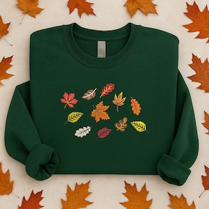 Puede incluir: Una sudadera verde oscuro con un diseño bordado de hojas de otoño coloridas. Las hojas son de color rojo, naranja, amarillo y blanco. La sudadera tiene cuello redondo y mangas largas. El fondo es de color beige claro con hojas de otoño dispersas.