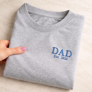 May include: A folded gray t-shirt with the word "DAD" embroidered in blue, along with "Est. 2026". The shirt is made of a soft material and has a crew neck.