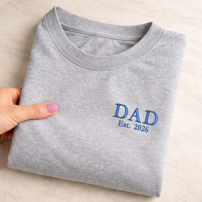 May include: A folded gray t-shirt with the word "DAD" embroidered in blue, along with "Est. 2026". The shirt is made of a soft material and has a crew neck.