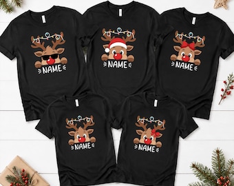 Könnte beinhalten: Fünf schwarze Kurzarm-T-Shirts mit einem Cartoon-Rentier-Motiv mit roter Nase und Lichterkette am Geweih. Jedes Shirt zeigt den Schriftzug NAME unter dem Rentier. Die Rentier-Designs variieren mit Sonnenbrille, Weihnachtsmütze oder einer roten Schleife.