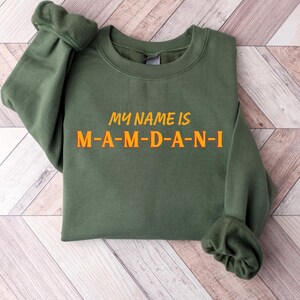 May include: Olive green sweatshirt with the text "MY NAME IS M-A-M-D-A-N-I" in orange. The sweatshirt is folded, with the sleeves visible. The background is a light wood grain pattern.