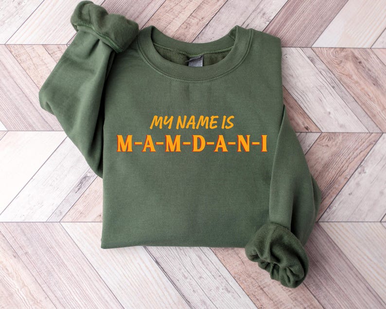 May include: Olive green sweatshirt with the text "MY NAME IS M-A-M-D-A-N-I" in orange. The sweatshirt is folded, with the sleeves visible. The background is a light wood grain pattern.