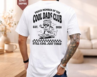 Könnte beinhalten: Ein weißes T-Shirt mit einer schwarzen Grafik auf der Rückseite. Das Design zeigt einen Kaffeebecher auf einem Skateboard mit dem Text: PROUD MEMBER OF THE COOL DADS CLUB, EST. 2026 und STILL COOL JUST TIRED. Ein Schachbrettmuster ist unter der Grafik.