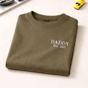 May include: Olive green crew neck sweatshirt with the embroidered text "DADDY Est. 2021" in white. The folded sweatshirt is on a light surface, with a toy car in the upper right corner.