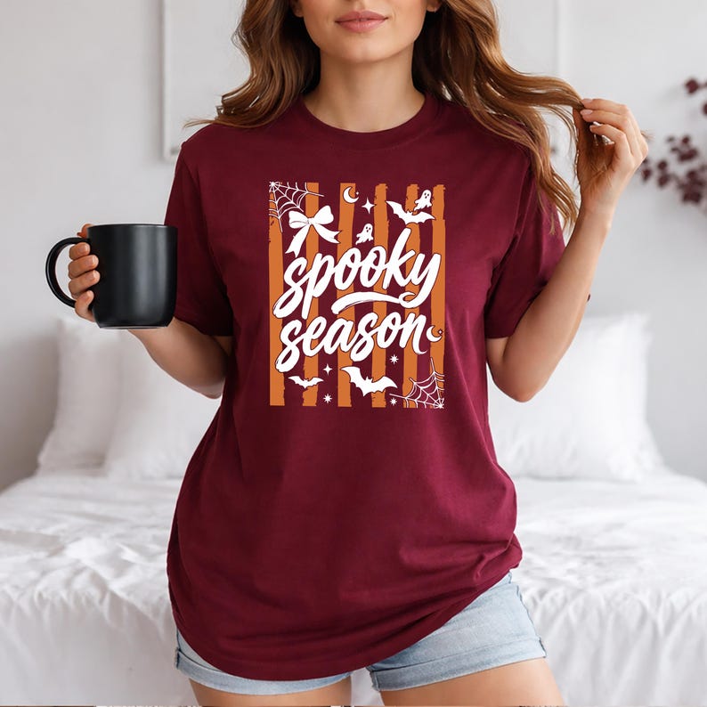 May include: A maroon short-sleeve t-shirt featuring a white graphic print with the text "spooky season" surrounded by illustrations of bats, spiderwebs, ghosts, and a bow, set against a background of vertical orange and maroon stripes.