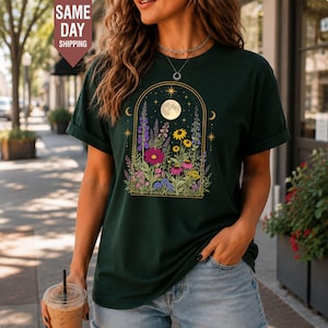 Op de afbeelding: Een donkergroen T-shirt met korte mouwen en een grafische print van een gouden boogvormig frame met een volle maan, sterren en een verscheidenheid aan kleurrijke wilde bloemen, waaronder paarse, roze en gele bloesems.