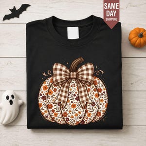 May include: A black crew neck t-shirt featuring a large graphic of a pumpkin with a brown and white gingham bow. The pumpkin is decorated with a pattern of small orange, brown, and cream-coloured flowers and leaves on a white background.