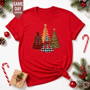 Może przedstawiać: Jaskrawoczerwony t-shirt z krótkim rękawem z grafiką przedstawiającą cztery stylizowane choinki. Drzewka mają różne wzory, w tym czerwoną kratę, złoty kolor, czarno-białą kratkę gingham oraz brązowy wzór w panterkę, a każde z nich zwieńczone jest gwiazdą.