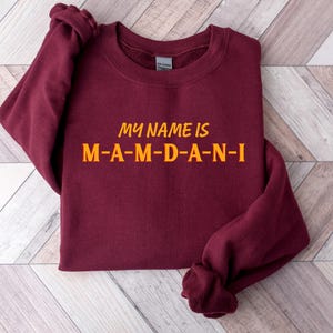 May include: A maroon sweatshirt with the text "MY NAME IS M-A-M-D-A-N-I" in orange lettering. The sweatshirt is folded, with the sleeves visible. The text is centered on the front of the garment.