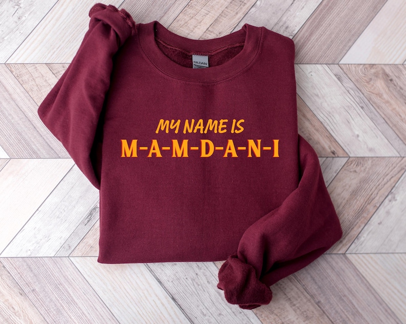 May include: A maroon sweatshirt with the text "MY NAME IS M-A-M-D-A-N-I" in orange lettering. The sweatshirt is folded, with the sleeves visible. The text is centered on the front of the garment.
