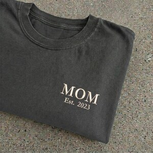May include: Dark gray crew neck t-shirt with the word "MOM" and "Est. 2023" printed in a light beige color. The shirt is folded, showing the text on the lower right side.
