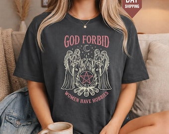 May include: A dark grey short-sleeve t-shirt featuring a graphic print of four figures standing around a campfire with a pentagram symbol. The text reads GOD FORBID WOMEN HAVE HOBBIES in pink lettering above and below the central illustration.