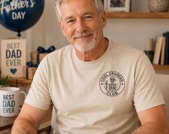 Könnte beinhalten: Beiges T-Shirt mit schwarzem "COOL GRANDAD CLUB"-Aufdruck, der ein Skelett mit Peace-Zeichen zeigt. Das Bild enthält auch einen "Happy Father's Day"-Ballon, eine "Best Dad Ever"-Karte und eine Tasse.