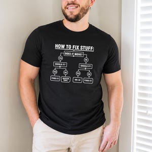 May include: A black short-sleeved t-shirt featuring a white graphic flowchart titled HOW TO FIX STUFF: with decision paths for Does it move? Should it? leading to outcomes including Fixed it!, Gaffer tape, and WD-40 in a clear, bold font.