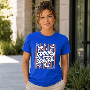 May include: A royal blue short-sleeve t-shirt featuring a graphic print with vertical orange stripes and white illustrations of spiderwebs, bats, stars, and a bow. The text "spooky season" is printed in a white script font across the centre.