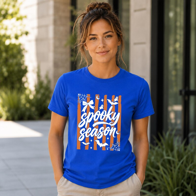 May include: A royal blue short-sleeve t-shirt featuring a graphic print with vertical orange stripes and white illustrations of spiderwebs, bats, stars, and a bow. The text "spooky season" is printed in a white script font across the centre.