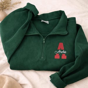 May include: A dark green sweatshirt with a quarter-zip neckline. The sweatshirt features a red letter "A" and the name "Archie" embroidered in white. The sleeves are folded, and the garment is lying on a cream-colored surface.