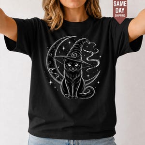 May include: A black short-sleeved t-shirt featuring a white graphic print of a black cat wearing a pointed witch hat, positioned in front of a crescent moon surrounded by small stars and swirling wisps of smoke or magical energy.