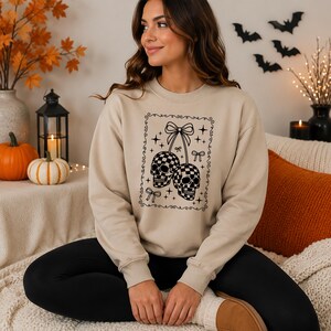 May include: A beige crewneck sweatshirt featuring a black graphic print of two checkered skulls hanging from a bow, surrounded by small stars and decorative bows within a rectangular border, with long sleeves and a ribbed collar and cuffs.