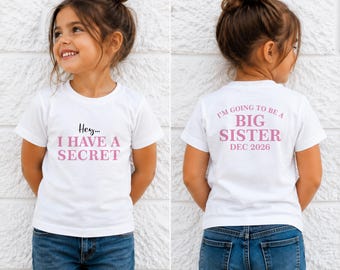 Könnte beinhalten: Ein weißes kurzärmeliges T-Shirt mit rosa Text. Die Vorderseite zeigt die Worte "Hey... I HAVE A SECRET" in Schwarz und Rosa. Die Rückseite trägt den Text "I'M GOING TO BE A BIG SISTER DEC 2026" in rosa Schrift auf dem weißen Stoff.