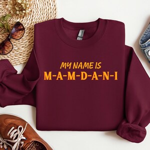 May include: A maroon sweatshirt with the text "MY NAME IS M-A-M-D-A-N-I" in yellow and orange lettering. The sweatshirt is displayed with sunglasses, brown shoes, and blue jeans.