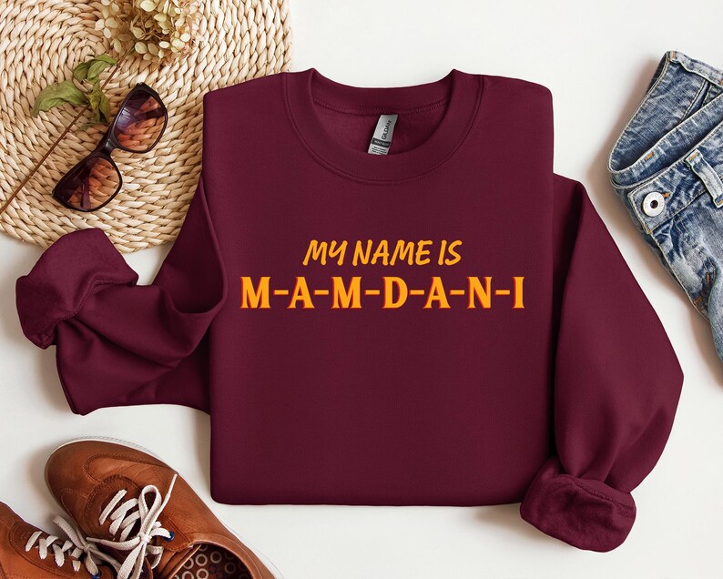 May include: A maroon sweatshirt with the text "MY NAME IS M-A-M-D-A-N-I" in yellow and orange lettering. The sweatshirt is displayed with sunglasses, brown shoes, and blue jeans.