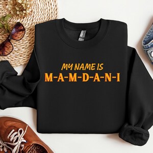 May include: Black sweatshirt with the text "MY NAME IS M-A-M-D-A-N-I" in orange and yellow lettering. The sweatshirt is displayed on a white surface with brown shoes, sunglasses, and denim jeans.