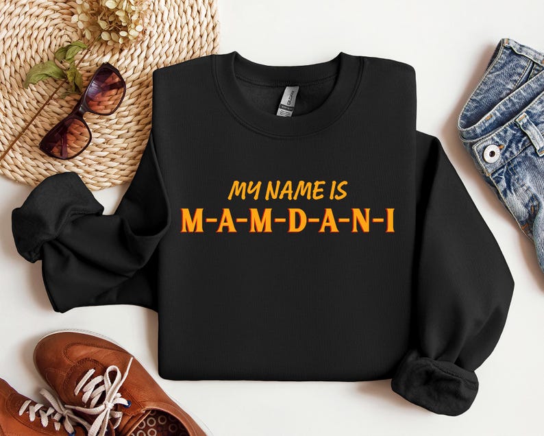 May include: Black sweatshirt with the text "MY NAME IS M-A-M-D-A-N-I" in orange and yellow lettering. The sweatshirt is displayed on a white surface with brown shoes, sunglasses, and denim jeans.