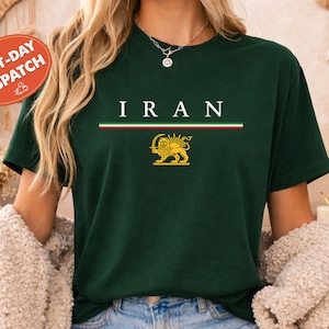 Puede incluir: Una camiseta verde oscuro con la palabra "IRAN" en letras blancas. Debajo, una franja horizontal en rojo, blanco y verde. Un emblema de león dorado está debajo. La camiseta es de cuello redondo y manga corta.