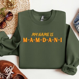 May include: An olive green sweatshirt with the text "MY NAME IS M-A-M-D-A-N-I" in orange and yellow. The sweatshirt is a crew neck style. The image also includes brown shoes, sunglasses, and denim jeans.