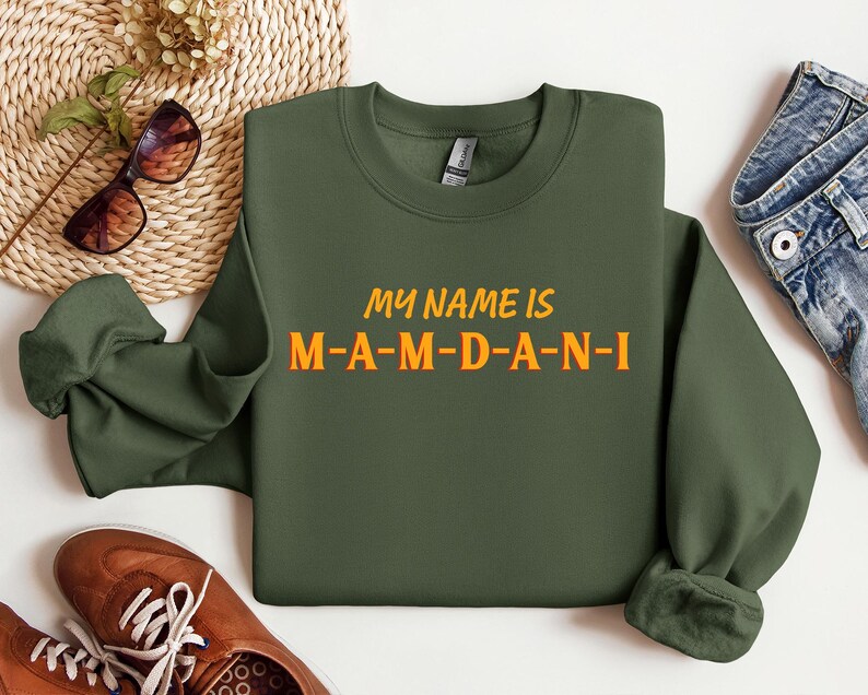 May include: An olive green sweatshirt with the text "MY NAME IS M-A-M-D-A-N-I" in orange and yellow. The sweatshirt is a crew neck style. The image also includes brown shoes, sunglasses, and denim jeans.