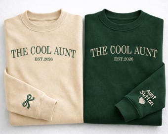 Könnte beinhalten: Zwei Sweatshirts, eines beige und eines dunkelgrün, mit der Stickerei "THE COOL AUNT EST.2026" auf der Vorderseite. Das beige Sweatshirt hat eine Schleife am Ärmel, das grüne sagt "Aunt Sutton" mit einem Herz.