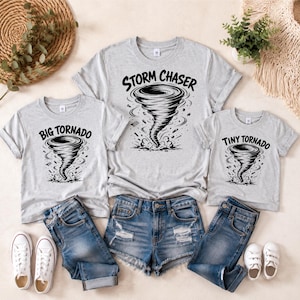 Puede incluir: Tres camisetas de color gris claro con estampados gráficos negros de tornados. La camiseta más grande muestra el texto "STORM CHASER", mientras que las dos más pequeñas presentan los textos "BIG TORNADO" y "TINY TORNADO" en negrita.