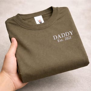 May include: Olive green crew neck t-shirt with the white text "DADDY Est. 2021" printed on the front. The shirt is folded neatly, showcasing the design. The shirt is made of a soft material.